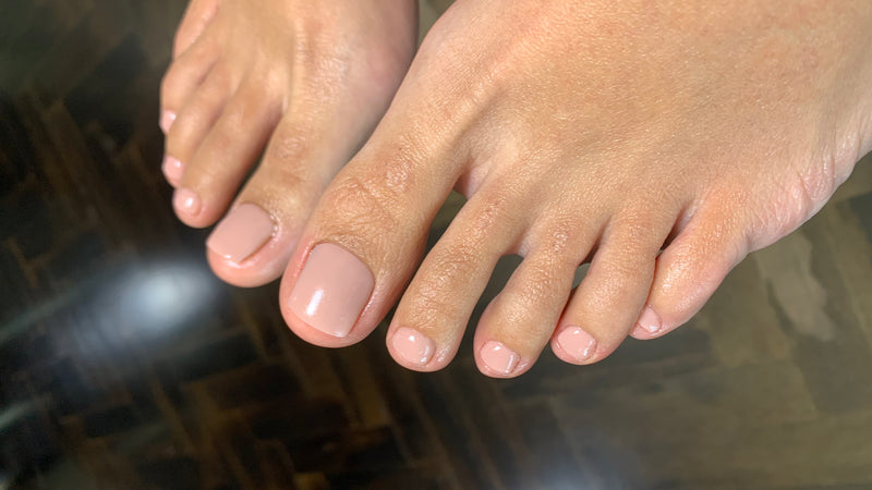 Close-up of a person's feet with neatly pedicured toenails painted in a light creamy nude shade, with dark wooden flooring in the background